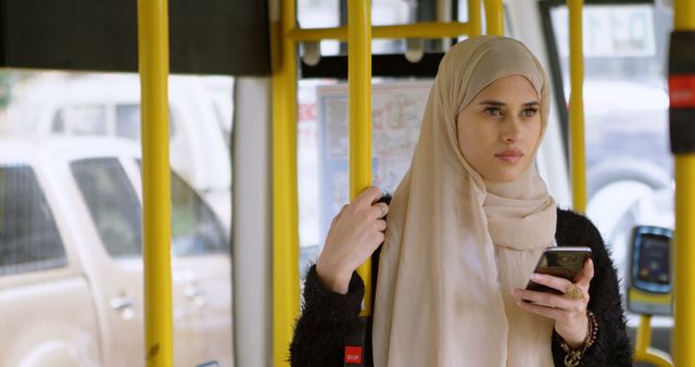 A young Muslim woman in hijab is commuting on a public bus while holding her smartphone. She appears thoughtful and focused, creating a scene relatable to daily public transportation users. This picture can be used in contexts related to public transportation, modern technology, urban lifestyle, diversity, and cultural representation.
