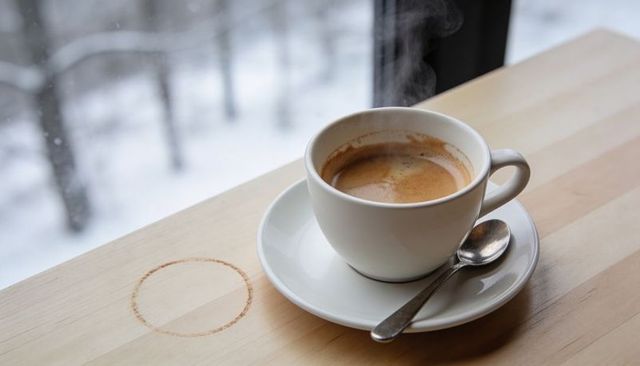 Steaming espresso cup on saucer with teaspoon and coffee ring on wooden table, snowy view