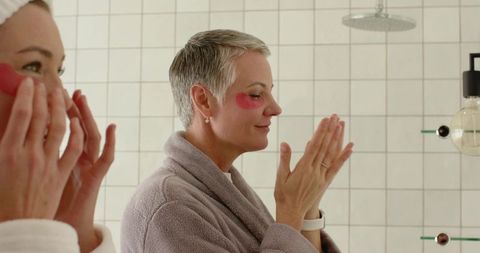 Mother and daughter applying pink under-eye patches at tiled bathroom mirror, self-care