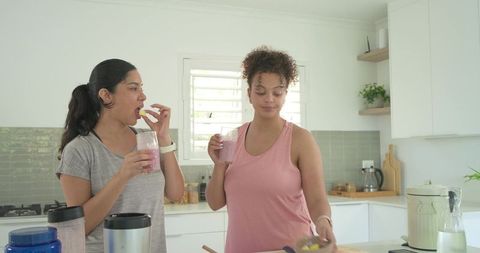 Two diverse women preparing and sipping pink smoothies in bright modern kitchen