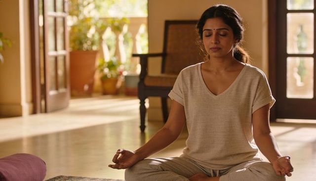 Meditating indian woman wearing loungewear, sitting cross-legged on sunlit rug by balcony