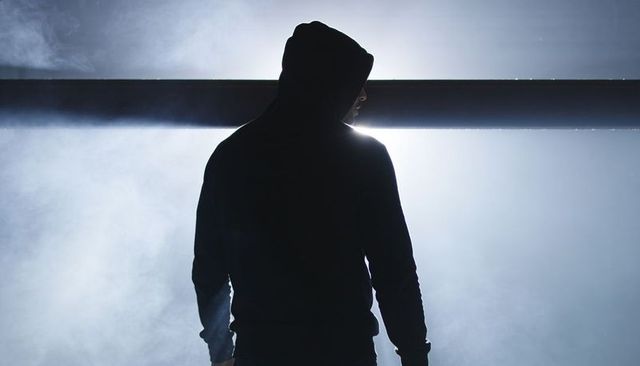 Hooded figure standing backlit in fog, looking right, dramatic metal beam halo