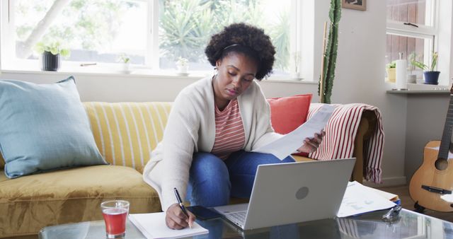 Woman sitting in living room with laptop and documents. She appears focused and productive in a home office setting. Ideal for illustrating concepts related to remote work, freelancing, and home office productivity.