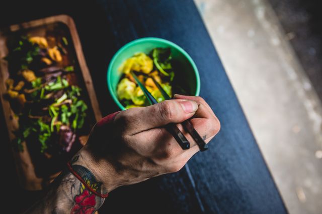 Close-up of a tattooed hand using chopsticks to eat fresh salad served in a green bowl. The setting conveys a healthy eating theme with bright colors and fresh ingredients. Ideal for use in health and lifestyle blogs, food websites, or advertisements promoting vegetarian or healthy meals.