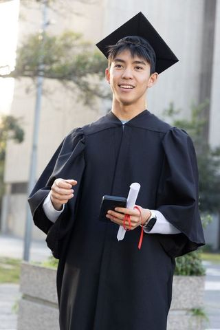 Young asian graduate celebrating on campus holding diploma and smartphone wearing mortarboard