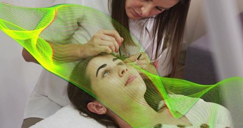 Esthetician shaping eyebrows with tweezers on reclining client with green mesh effect