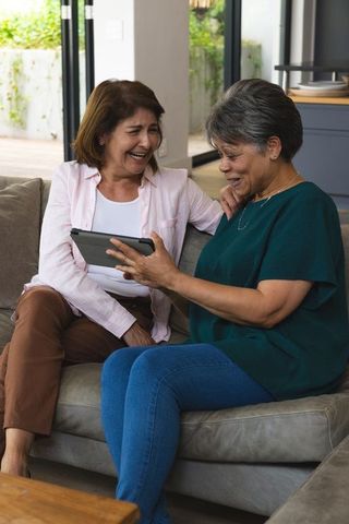 Senior Women Laughing Together While Using Tablet In Cozy Living Room