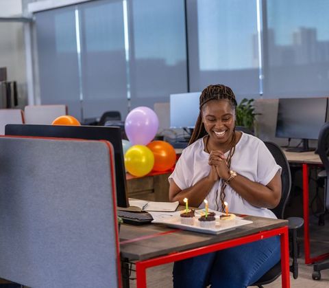 African american woman celebrating birthday at office desk with cupcakes and balloons