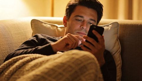 Man relaxing on couch tapping smartphone under cozy blanket in warm ambient light
