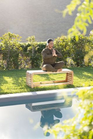 Asian woman meditating on bench by pool wearing headphones and olive loungewear