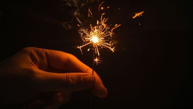 Hand holding lit sparkler at night, closeup showing bright golden festive sparks and embers