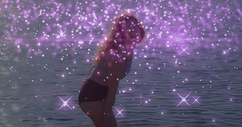 Young woman wading in sea wearing high-waisted bikini with purple sparkle bokeh overlay