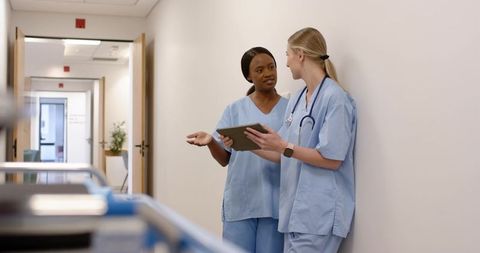 Two nurses discussing patient care in hospital corridor using tablet and stethoscope