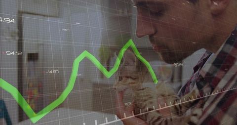 Mid-adult man cradling tabby kitten in home office with green financial chart overlay