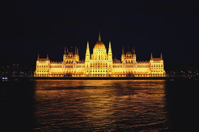 View of Hungarian Parliament Building illuminated at night alongside the calm waters of the Danube River. Perfect for travel blogs, European tourism promotions, and architectural competitions. Can be used in content showcasing iconic landmarks of Budapest and night photography.