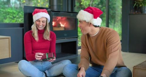 Couple exchanging holiday gifts by fireplace wearing santa hats in modern living room
