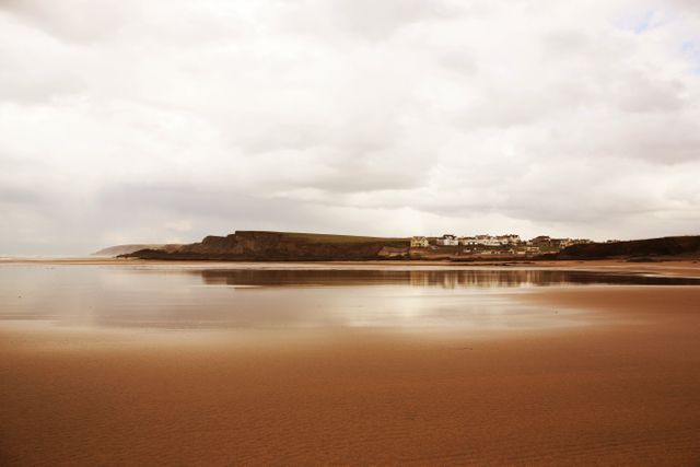 Tranquil coastal scene features empty beach, distant houses, and cliffs under a cloudy sky. Reflections on wet sand enhance serene atmosphere. Ideal for relaxation themes, marketing for coastal properties, travel posters, or inspirational content. Use for blogs, websites, flyers promoting beach destinations or real estate catalogues.