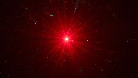 Radiating red starburst with radial beams and teal particle flecks in dark void