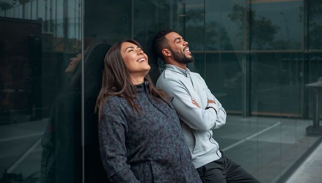 Laughter break between coworkers leaning against glass facade, relaxed urban teamwork