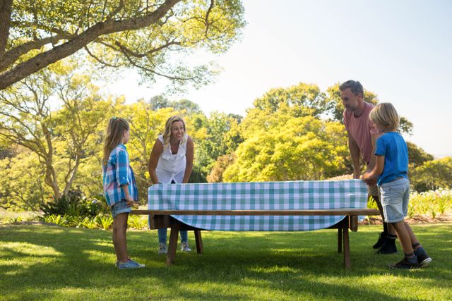 Family spreading tablecloth on picnic table in a park, surrounded by lush greenery. Ideal for use in advertisements, articles, and blogs about family activities, outdoor fun, and summer picnics. Perfect for promoting family bonding, leisure activities, and nature outings.