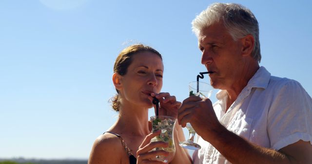 A middle-aged Caucasian couple enjoys a refreshing drink together under a clear blue sky, with copy space. Their shared moment reflects a relaxed and intimate connection, during a vacation or a leisurely day out.