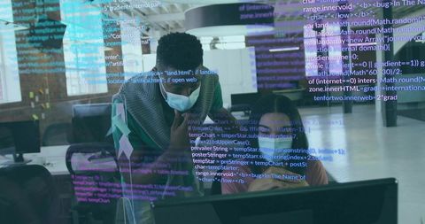 Masked woman leaning over colleague guiding coding and debugging on monitor in open-plan office