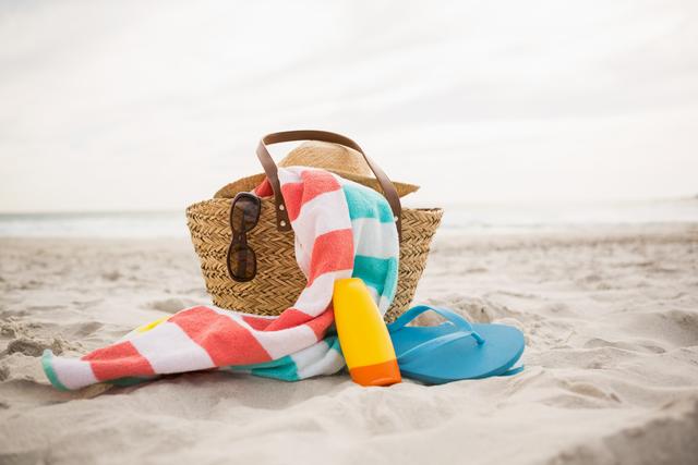 Colorful beach accessories including a striped towel, sunglasses, a hat, blue flip flops, and sunscreen on sandy shore with sea in background. Perfect for vacation, travel, summer promotions, touristic advertisements, and relaxation-themed projects.