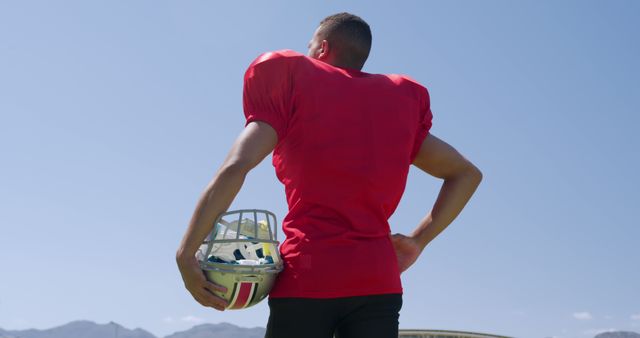 Football player wearing red jersey stands outdoors holding his helmet. Ideal for sports promotions, athletic training materials, football event advertisements, and stock imagery for articles about sportsmanship.