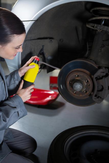 Female mechanic oiling a car brake in an auto repair shop. This image can be used in automotive repair advertisements, articles about women in trades, and educational materials on vehicle maintenance.
