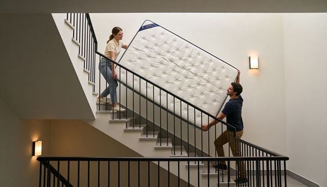 Couple carrying tufted mattress up modern staircase showing teamwork during move