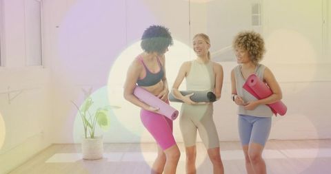 Diverse women holding yoga mats laughing in bright fitness studio after class