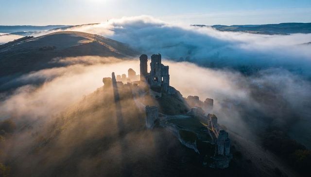 Medieval castle ruins bathing in golden sunrise mist over hill ridge aerial landscape