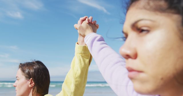 Two women holding hands with blue sky and ocean in the background. Suggests themes of friendship, unity, and support. Ideal for use in advertisements, blogs, or social media promoting friendship, outdoor activities, or wellness.