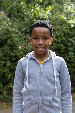 African american boy standing in garden wearing gray zip-up hoodie smiling in sunlight