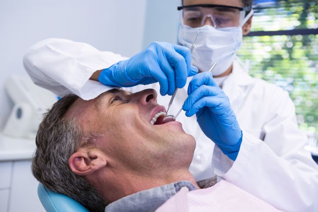 Man receiving dental treatment from a dentist in a clinic. Dentist wearing protective gear and using dental tools. Ideal for illustrating dental care, oral health, medical services, and healthcare advertisements.