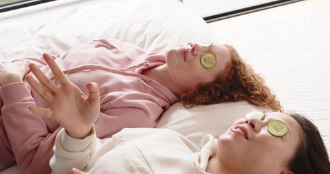 Two friends enjoying diy home spa day with cucumber eye slices relaxing on bed