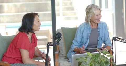 Two senior women recording podcast in sunlit living room, collaborating with tablet and mics
