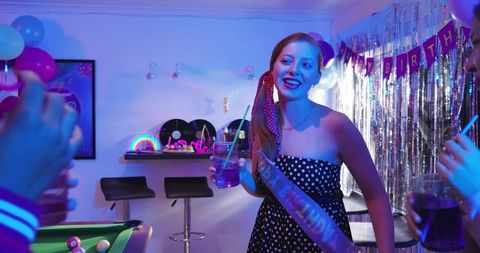 Woman wearing sash enjoying neon birthday party with friends at pool table holding purple cocktail