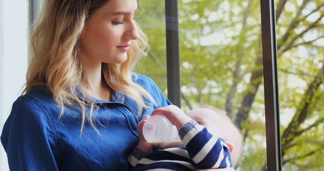 Young mother feeding baby with bottle by window - Download Free Stock Images Pikwizard.com