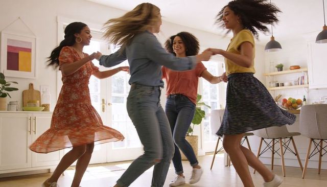 Four friends dancing in sunlit kitchen holding hands, spinning joyful circle
