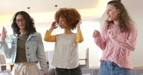 Multicultural young women dancing laughing singing together in bright modern living room