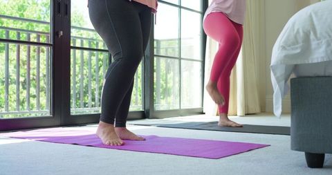 Female friends practicing balance on yoga mats in sunlit bedroom with balcony view