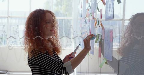 Red-haired woman placing sticky notes on glass wall during creative brainstorming session