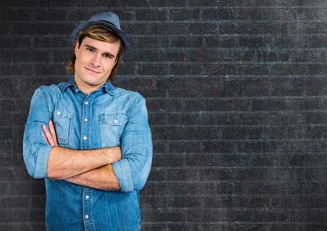 Young man wearing a hat and denim shirt, standing confidently with crossed arms against a dark brick wall background. Useful for advertising, lifestyle blogs, fashion editorials, or promotional materials focusing on youth, style, or casual fashion trends.