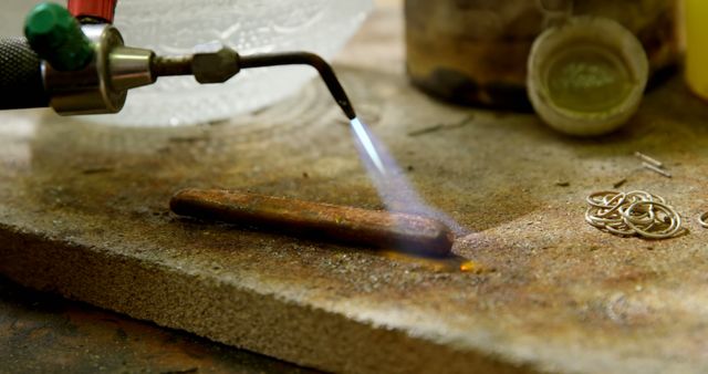 A jeweler's torch is heating a metal rod on a workbench, with tools and materials indicative of jewelry making in the background. This close-up captures the precision required in the craftsmanship of jewelry, with the flame's heat essential for shaping and soldering metal components.