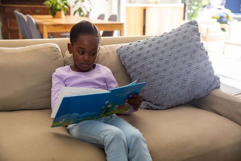 School-age african american girl reading colorful storybook on cozy living room sofa