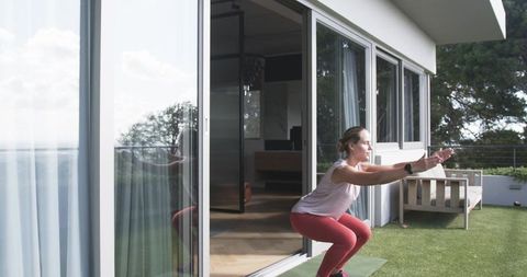 Woman performing outdoor squats on patio mat near sliding glass doors overlooking garden