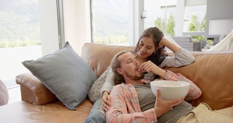 Couple enjoys relaxing on a comfortable sofa at home, sharing a fun moment with popcorn while watching a movie. Ideal for themes related to home comfort, relationships, leisure activities, and lifestyle blogs. This scene showcases warmth and togetherness, perfect for marketing cozy home environments or snack products.
