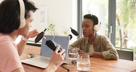 Two diverse coworkers recording conversational podcast with laptop and microphones in sunlit studio