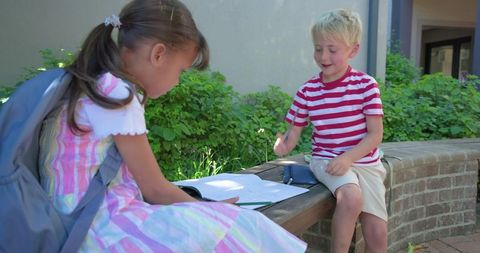 Children studying together on school bench in sunny courtyard sharing notebook and pencil